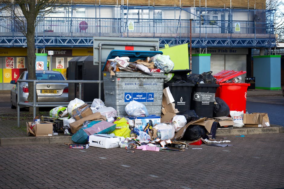 Close-up view of a large green waste skip positioned against a dark brick wall in an outdoor setting. The skip is filled with black and red bags of building or garden waste, some of which are branded