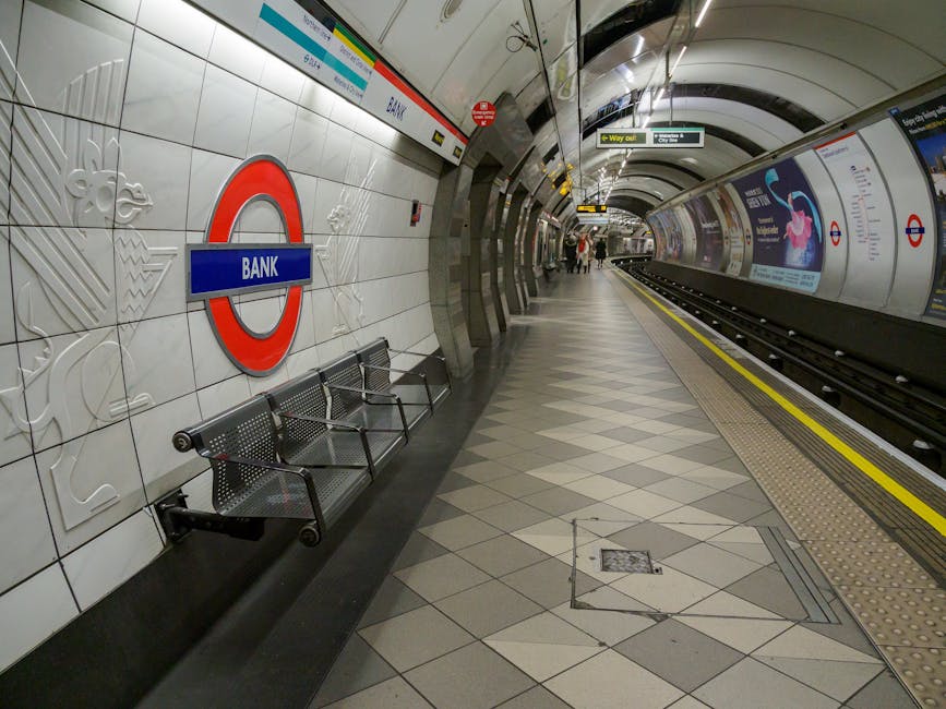 The open doors of a red London Underground train are visible at a station platform, with the interior showing two rows of seats facing forwards, a blue handrail, and safety signage near the doorway. T