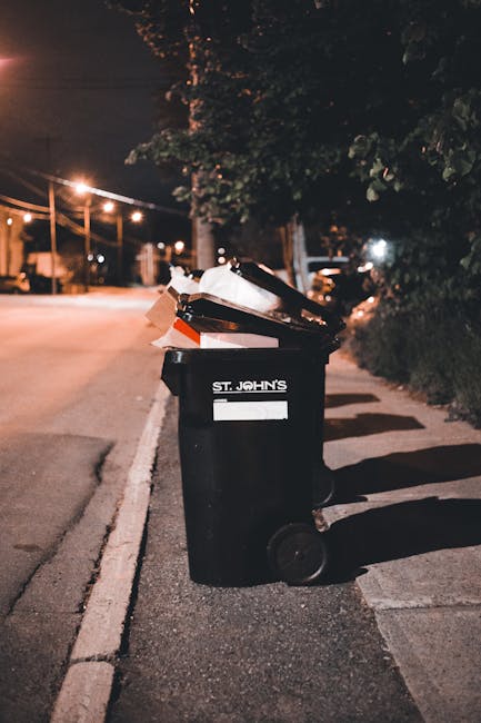 A large green waste disposal bin filled with various types of rubbish, including flattened cardboard boxes, small open cartons, and packaging materials, is positioned on a city street. The cardboard b